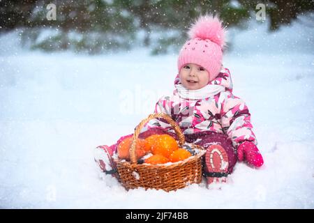 Una bambina in abiti invernali è seduta sulla neve con un cestino di arance. Foto Stock