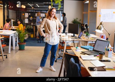 Ritratto di sorridente caucasica giovane donna d'affari in piedi con le mani sui fianchi sul posto di lavoro Foto Stock
