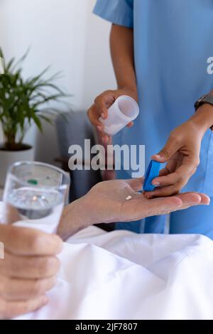 Metà sezione di lavoratore di salute femminile biraciale che dà pillole mediche per l'uomo anziano caucasico a letto Foto Stock