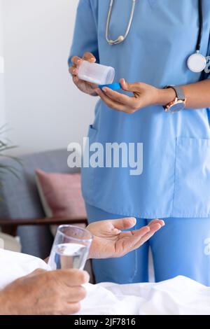 Sezione centrale dell'operatrice di salute femminile di biracial che dà le pillole mediche all'uomo anziano che si trova sul letto Foto Stock