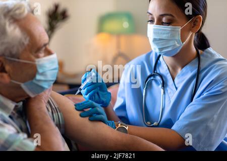 Professionista della salute femminile biracale che fa un'iniezione all'anziano caucasico a casa Foto Stock