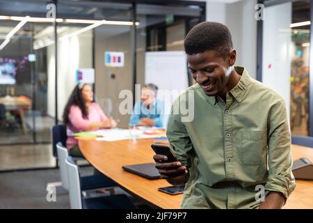 Sorridendo il giovane uomo d'affari afroamericano usando lo smart phone mentre i colleghi in background Foto Stock