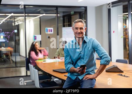 Ritratto di uomo d'affari caucasico maturo sorridente con smartphone mentre colleghi in background Foto Stock