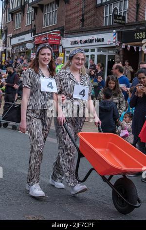 Due donne in abito fantasia spingono una carropola giù Pinner High Street prima della corsa carropola alla festa di St Georges' Day Celebration. Pinner, NW Londra. Foto Stock