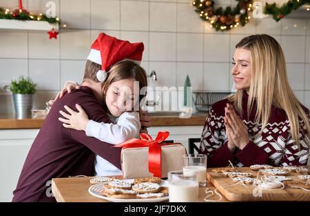 Papà a santa cappello che presenta il regalo di Natale alla figlia del bambino che abbraccia a casa. Foto Stock
