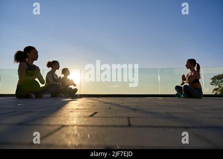 Diverse silhouette femminili e istruttore di yoga seduti all'aperto meditating. Foto Stock