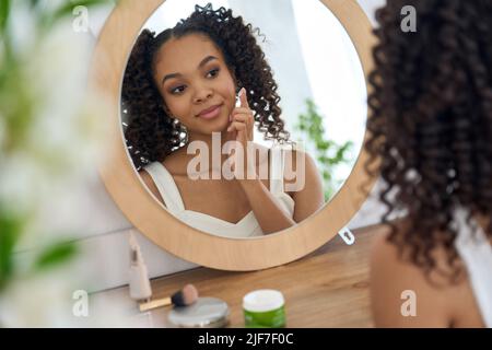 Ragazza africana teen guardando nello specchio del bagno che applica la crema facciale sul viso. Foto Stock