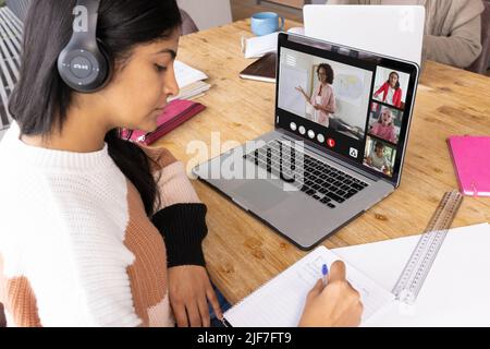 Ragazza adolescente biraciale indossando cuffie scrittura mentre segue professore insegnamento in classe online Foto Stock