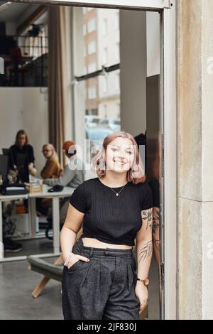 Imprenditore sorridente femminile con mano in tasca appoggiata sulla porta d'ufficio Foto Stock