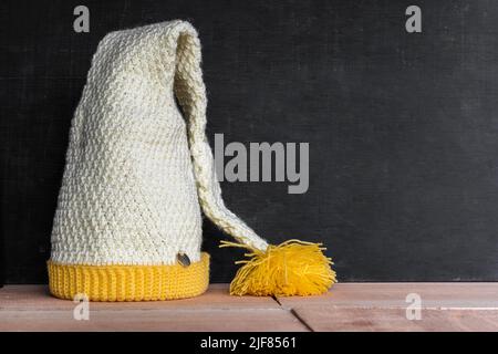 cappello di elfo a crochet, posto su sfondo di legno nero e arancione, rifinito e pronto per essere utilizzato nel duro inverno. cappello di bambino giallo e bianco. spazio Foto Stock