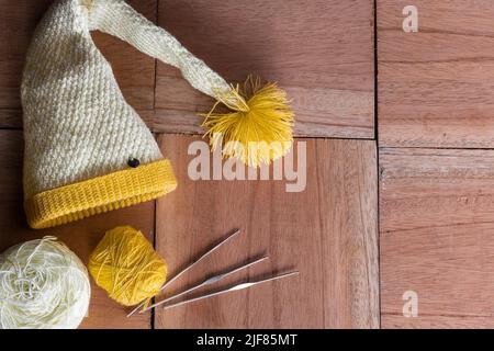 ancora vita di giallo e bianco elfo cappello per l'inverno, a mano lavorato a crochet su uno sfondo di legno colore arancione, accanto ad esso due rotoli di lana e tre Foto Stock