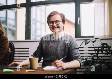 Ritratto di giovane imprenditore maschio felice indossando occhiali mentre si siede al tavolo di conferenza Foto Stock