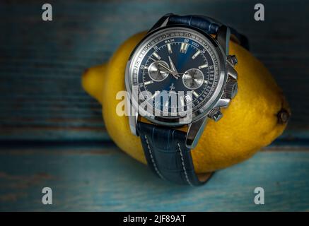 Studio girato di un elegante orologio da polso blu uomo di lusso avvolto intorno al limone giallo, su un tabke di legno blu. Accessori per uomo in stile business. Foto Stock