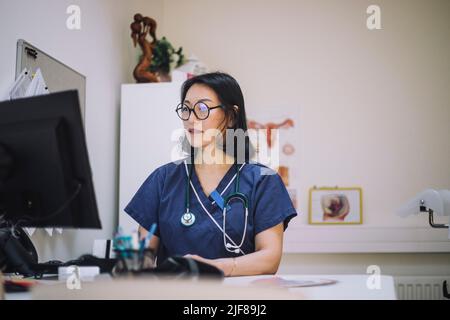 Medico femminile maturo che indossa occhiali utilizzando il computer seduto alla scrivania in ospedale Foto Stock