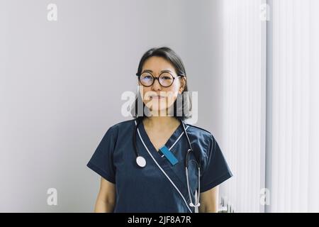 Ritratto di medico femminile che indossa occhiali in piedi contro la parete bianca in clinica medica Foto Stock
