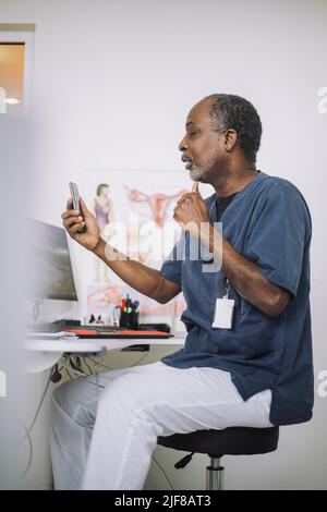 Medico maschile che fa la consultazione online attraverso il telefono cellulare mentre si siede alla scrivania in clinica Foto Stock