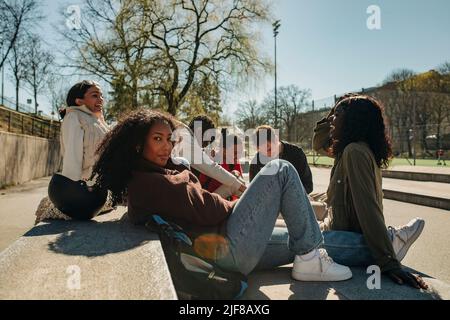 Ritratto della vista laterale della donna appoggiata sul muro di ritegno da amici multirazziali in parco il giorno di sole Foto Stock