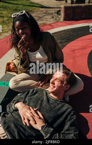 Uomo sorridente sdraiato sul grembo di un'amica mentre si rilassa nel parco giochi nelle giornate di sole Foto Stock