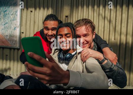 Sorridenti amici maschi multirazziali che fanno videochiamata attraverso smartphone contro muro Foto Stock