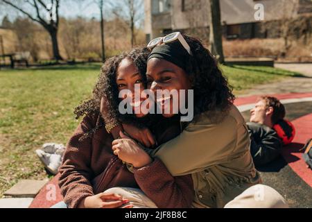 Felici amici multirazziale femmina abbracciarsi mentre si siede nel parco giochi in giorno di sole Foto Stock