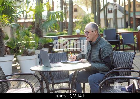 Vista laterale dell'uomo di mezza età che indossa una giacca nera, seduta al tavolo in un bar, scrittura di note in blocco appunti guardando il computer portatile. Foto Stock