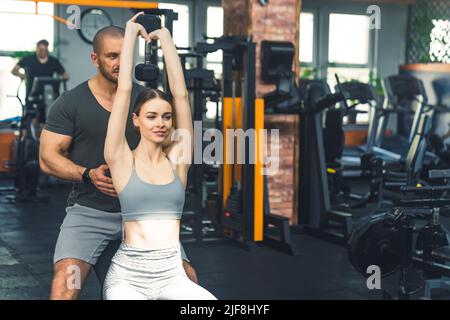 Bella donna sottile dai capelli scuri nel suo 20s seduto su una panca e sollevando un pesante dumbbell con la schiena dritta. Allenatore personale maschile in piedi dietro di lei e aiutare. Foto di alta qualità Foto Stock