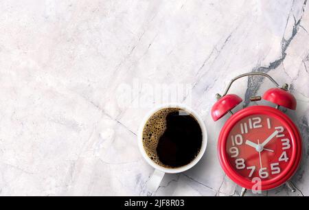 Caffè nero mattutino in tazza bianca e sveglia rossa su sfondo in marmo bianco. Vista dall'alto con spazio per la copia. Foto Stock