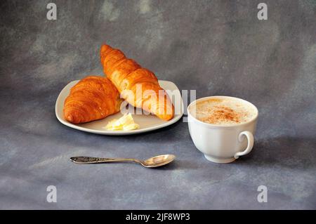 Due croissant freschi con burro su un piatto e una tazza di cappuccino caldo con cannella su sfondo grigio. Primo piano. Foto Stock