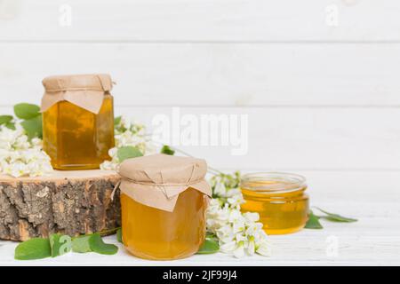 Dolce vaso di miele circondato primavera fiori di acacia. Il miele scorre da un cucchiaio in un vaso. Vasetti di miele di acacia fresco chiaro su sfondo di legno. Foto Stock