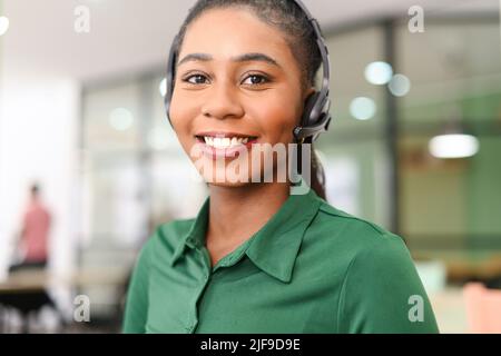 Headshot di giovane dipendente afroamericano che indossa cuffie wireless, guardando la fotocamera con un sorriso. Donna allegra in cuffie con microfono, supporto tecnico in ufficio Foto Stock