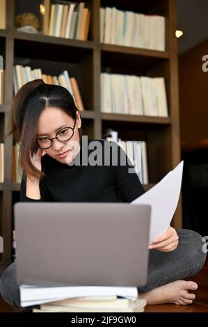 Focalizzato giovane donna asiatica freelancer o scrittore concentrato leggere alcuni contenuti online sullo schermo del laptop, lavorando nel suo ufficio domestico. Foto Stock