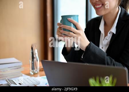 Una giovane donna d'affari asiatica allegra o una lavoratrice di ufficio femminile in abito formale sorseggiando un caffè alla sua scrivania. Immagine ritagliata Foto Stock