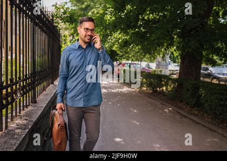 Uomo d'affari sicuro che discute sullo smartphone contro gli alberi. Il pendolari dirigente maschile cammina accanto alla recinzione sul marciapiede. Indossa i formals nella c Foto Stock