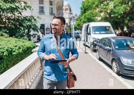 Un imprenditore sorridente che utilizza un tablet digitale. I pendolari maschili dell'esecutivo guardano lontano mentre camminano sul marciapiede. Indossa i formals in città durante Foto Stock
