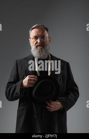 uomo anziano bearded in cappotto ed occhiali eleganti che tengono il cappello di derby sul grigio Foto Stock