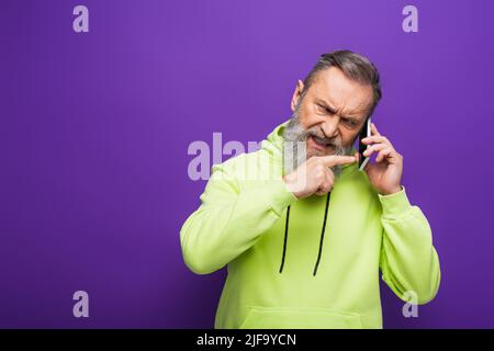 uomo anziano arrabbiato con cappuccio verde che punta con un dito mentre parla sullo smartphone su viola Foto Stock