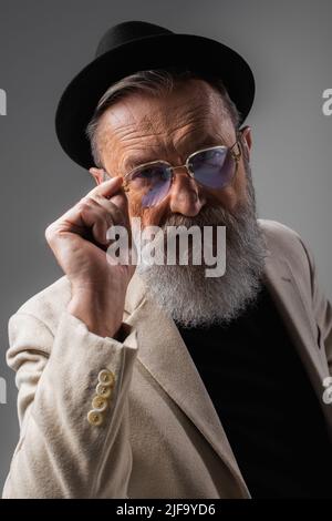 elegante uomo anziano in giacca beige e cappello da derby che regola gli occhiali sul grigio Foto Stock