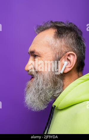 vista laterale dell'uomo anziano felice con cappuccio verde che ascolta musica in auricolari wireless viola Foto Stock