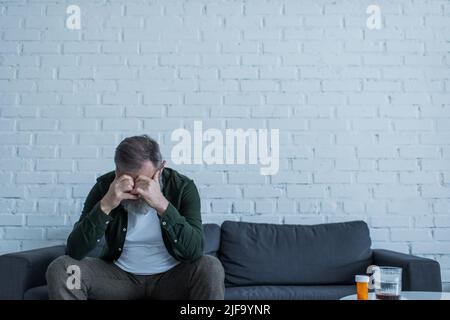 uomo anziano depresso con capelli grigi seduti sul divano accanto alla bottiglia con medicazione e bicchiere di whiskey sul tavolino da caffè Foto Stock