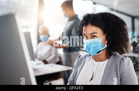 Agente di telemarketing misto per call center che indossa la maschera facciale come protocollo di sicurezza e parla con un auricolare mentre utilizzi il computer in ufficio. Femmina Foto Stock