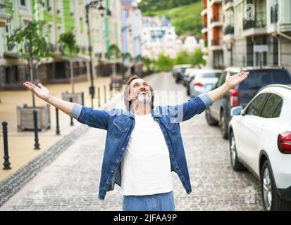 Felice uomo anziano grazie a Dio in piedi all'aperto con le mani spalancate ampia camicia da indossare jeans con t-shirt bianca sotto. Uomo di mezza età contento che la vita è bella in piedi nella strada della città vecchia. Foto Stock