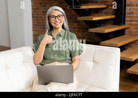 Vista frontale di una donna anziana e rilassata freelancer, comodamente seduta sul divano nel soggiorno, mostrando un dito grande, blogging, lavorando sul progetto da casa Foto Stock