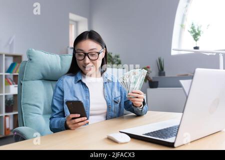 La donna di affari asiatica riuscita è felice con vincita nel casino in linea, guardando lo schermo del telefono e tenendo i dollari dei soldi nelle mani, wor della donna di affari Foto Stock