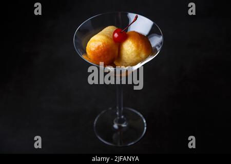 Rum baba napoletano (o baba au rum) in un bicchiere di martini con una ciliegina da cocktail su sfondo nero. Piccole torte di lievito imbevute di sciroppo di rum. Tradizione Foto Stock