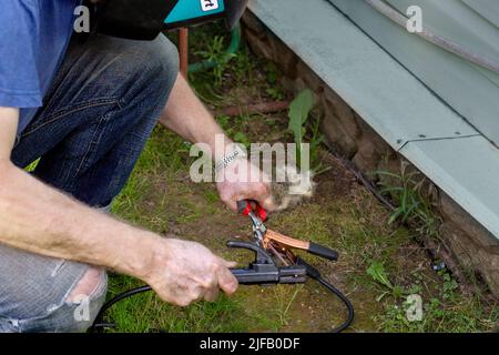 Le mani di un uomo tengono una saldatrice e sono impegnate nella saldatura di metallo e riparazione di una porta di auto sullo sfondo di una casa di campagna. Uomini t Foto Stock