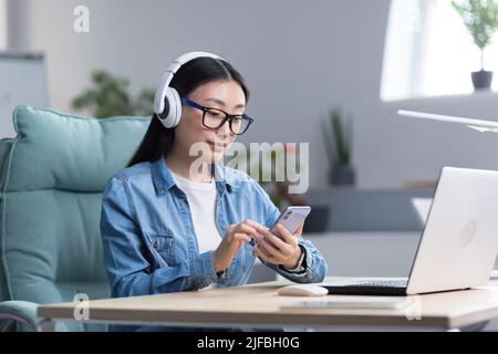 Giovane donna asiatica che ascolta la musica in ufficio, utilizzando il telefono e l'app musicale, donna d'affari con cuffie che riposano. Foto Stock