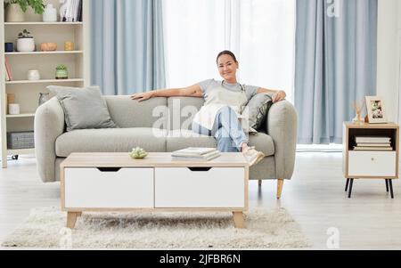 Una felice sorridente donna da corsa mista che si rilassa sul divano dopo aver pulito il suo appartamento. Una donna asiatica seduta sul divano e dopo una pausa Foto Stock