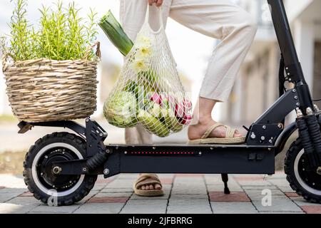 Donna guida su scooter elettrico con sacco a rete pieno di verdure, primo piano Foto Stock