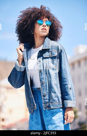 Donna ispanica alla moda e alla moda che indossa occhiali da sole all'esterno. Allegra giovane donna da corsa mista con un riccio afro che indossa un denim alla moda ed elegante Foto Stock