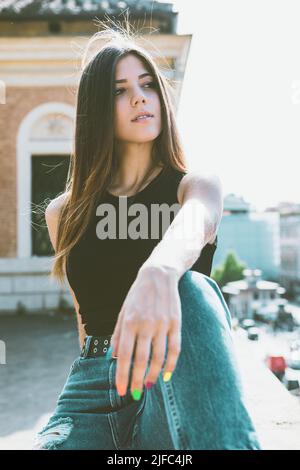 Bella ragazza seduta in città. Abito e stile urbano, jeans e top nero. Smalto per unghie colorato. Luce rilassata e luminosa. Foto Stock